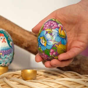 May include: A hand holds a purple Easter egg decorated with colourful flowers and green leaves. The egg is sitting on a woven basket.