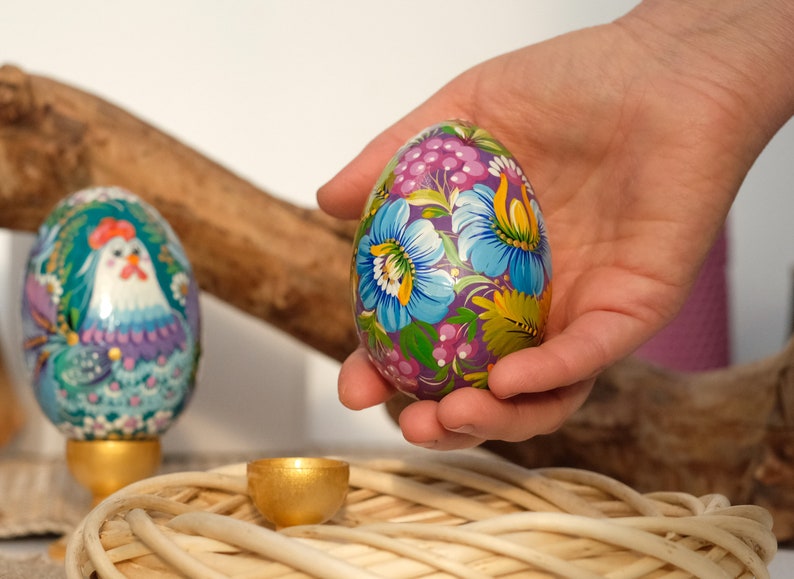 May include: A hand holds a purple Easter egg decorated with colourful flowers and green leaves. The egg is sitting on a woven basket.
