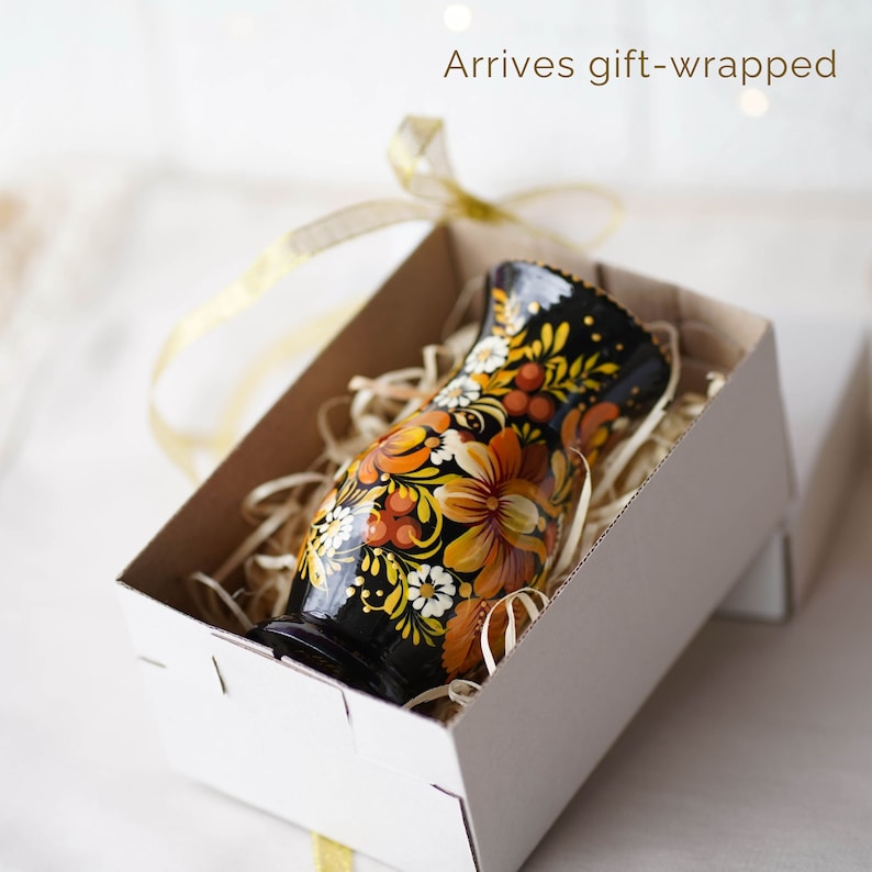 May include: A small black vase with a floral design in shades of orange, yellow, and white. The vase is in a white gift box with shredded paper.