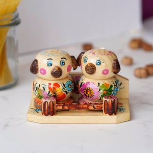 May include: A set of wooden salt and pepper shakers shaped like dogs with painted floral designs. The shakers are sitting on a wooden stand.