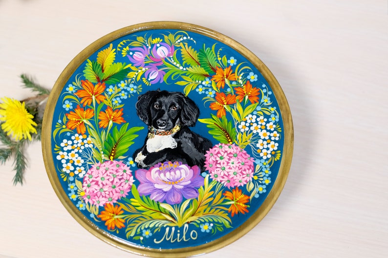 Puede incluir: Un plato redondo de madera con fondo azul y un dise&ntilde;o floral pintado. Un perro blanco y negro est&aacute; en el centro del plato, rodeado de flores. El nombre del perro, "Milo", est&aacute; escrito en cursiva en la parte inferior del plato.