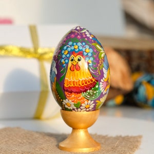 May include: A hand-painted Easter egg with a purple background and a colourful rooster design. The egg is displayed on a gold stand.