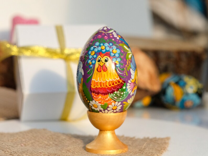 May include: A hand-painted Easter egg with a purple background and a colourful rooster design. The egg is displayed on a gold stand.