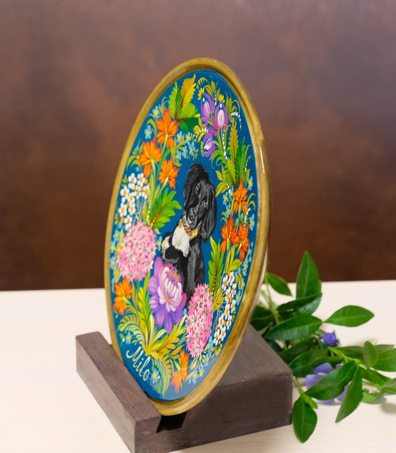 Puede incluir: Un plato redondo de madera con fondo azul y un perro blanco y negro pintado en el centro. El plato est&aacute; decorado con flores de colores y el nombre "Milo" est&aacute; pintado en la parte inferior.