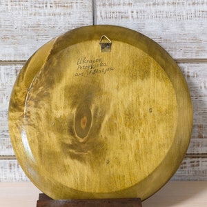 Puede incluir: La parte posterior de un plato redondo de madera con una mancha marr&oacute;n claro. El plato tiene un colgador de metal sujeto a la parte superior. El plato est&aacute; firmado con "Ukraine Petrykivka art of Slatynova".