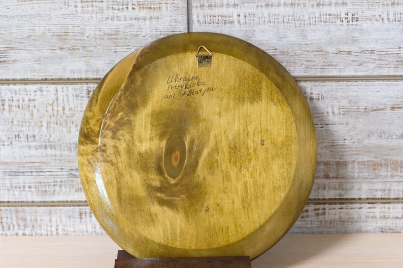 Puede incluir: La parte posterior de un plato redondo de madera con una mancha marr&oacute;n claro. El plato tiene un colgador de metal sujeto a la parte superior. El plato est&aacute; firmado con "Ukraine Petrykivka art of Slatynova".