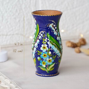 May include: A hand-painted wooden vase with a blue background and a floral design of white and blue flowers with green leaves. The vase is 4.7 inches (12 cm) tall.