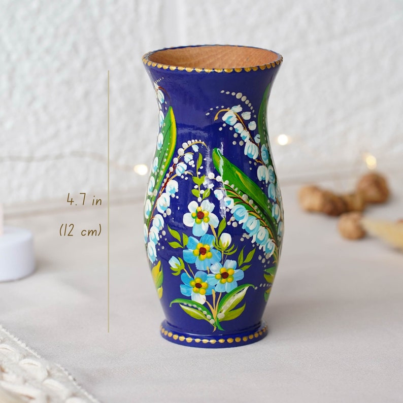 May include: A hand-painted wooden vase with a blue background and a floral design of white and blue flowers with green leaves. The vase is 4.7 inches (12 cm) tall.
