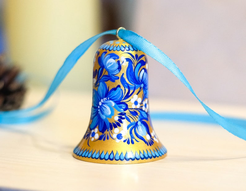 Puede incluir: Una campana dorada con dise&ntilde;os florales azules y blancos pintados en ella. La campana est&aacute; atada con una cinta azul claro.