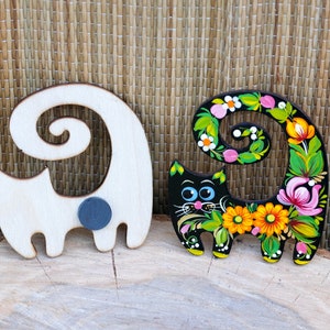 May include: Two wooden cat figurines, one painted black with colorful flowers and the other left natural wood. Both have a curled tail and a magnetic back.