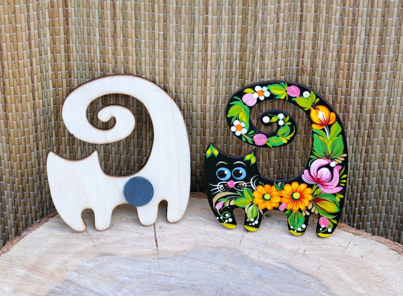 May include: Two wooden cat figurines, one painted black with colorful flowers and the other left natural wood. Both have a curled tail and a magnetic back.