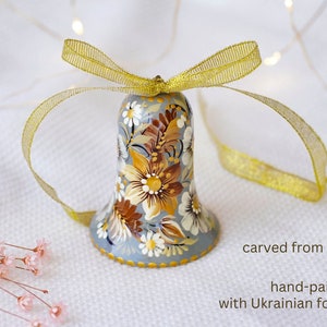 Op de afbeelding: Een handgeschilderd houten bel ornament versierd met een bloemenpatroon in tinten bruin, geel en wit op een grijze achtergrond. De bel is vastgebonden met een gouden lint en heeft de tekst "carved from wood hand-painted with Ukrainian folk art" eronder.