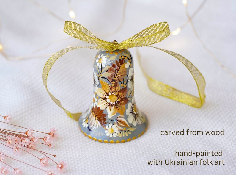 Op de afbeelding: Een handgeschilderd houten bel ornament versierd met een bloemenpatroon in tinten bruin, geel en wit op een grijze achtergrond. De bel is vastgebonden met een gouden lint en heeft de tekst "carved from wood hand-painted with Ukrainian folk art" eronder.
