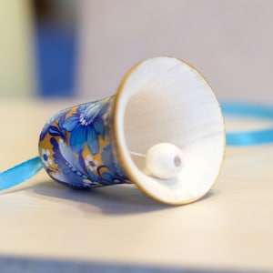 Puede incluir: Una peque&ntilde;a campana de madera pintada a mano con un dise&ntilde;o floral azul y dorado. La campana tiene un badajo blanco y est&aacute; atada con una cinta azul.