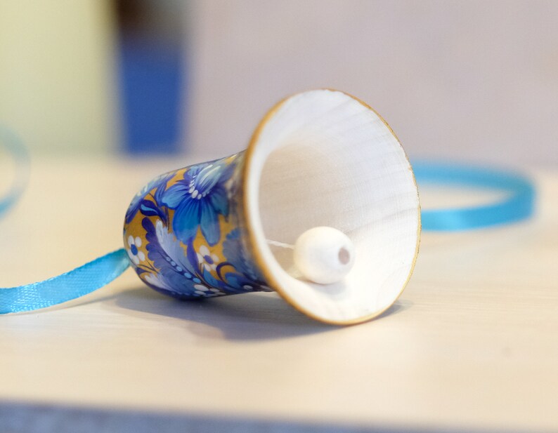 Puede incluir: Una peque&ntilde;a campana de madera pintada a mano con un dise&ntilde;o floral azul y dorado. La campana tiene un badajo blanco y est&aacute; atada con una cinta azul.