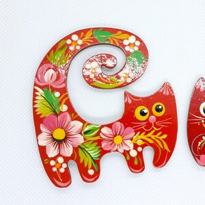 May include: A red wooden cat figurine with a curled tail, painted with pink and white flowers and green leaves. The cat has yellow eyes and a white smile.