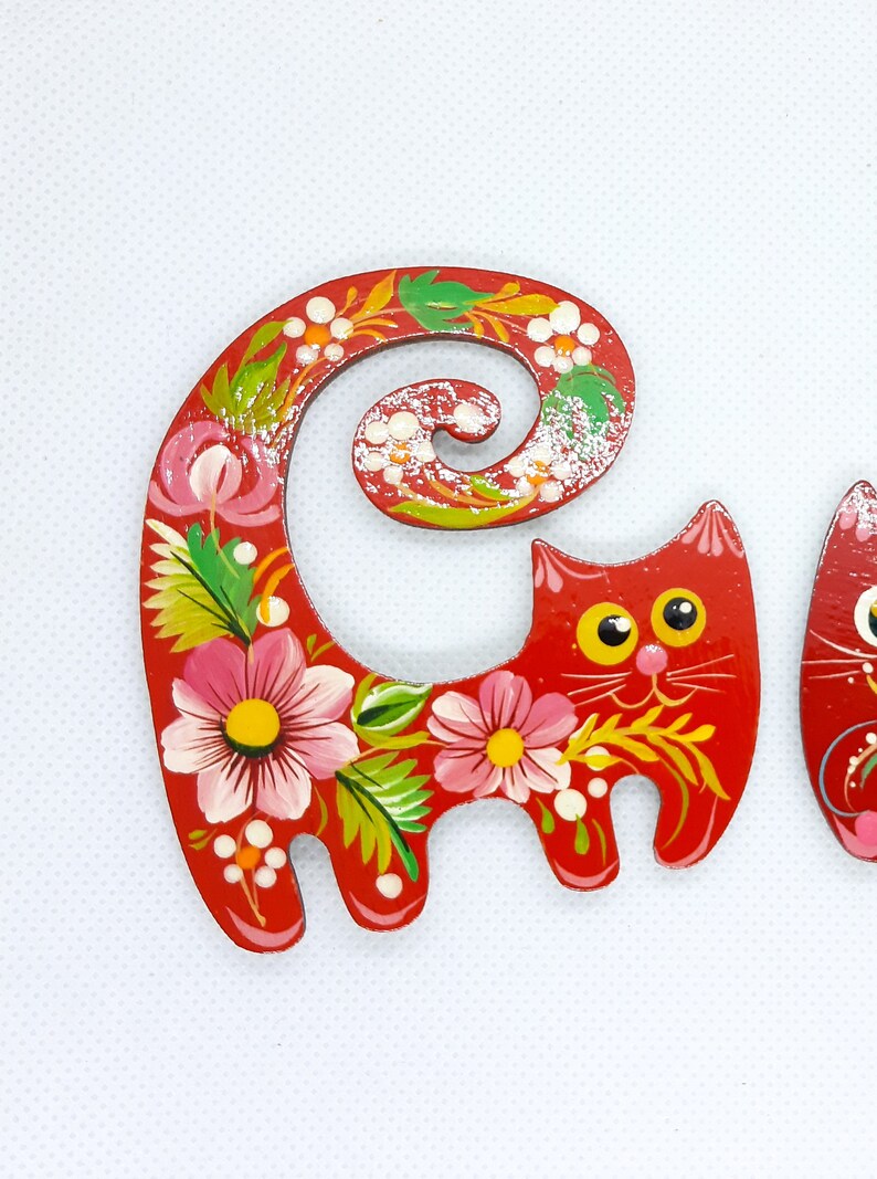 May include: A red wooden cat figurine with a curled tail, painted with pink and white flowers and green leaves. The cat has yellow eyes and a white smile.
