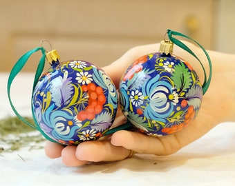 May include: Two blue and white hand-painted Christmas ornaments with floral designs and red berries. The ornaments are held in a hand.