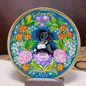 Puede incluir: Un plato de madera pintado a mano con un retrato de un perro blanco y negro rodeado de flores coloridas. El plato es azul con un borde dorado y el nombre "Milo" est&aacute; escrito en la parte inferior.