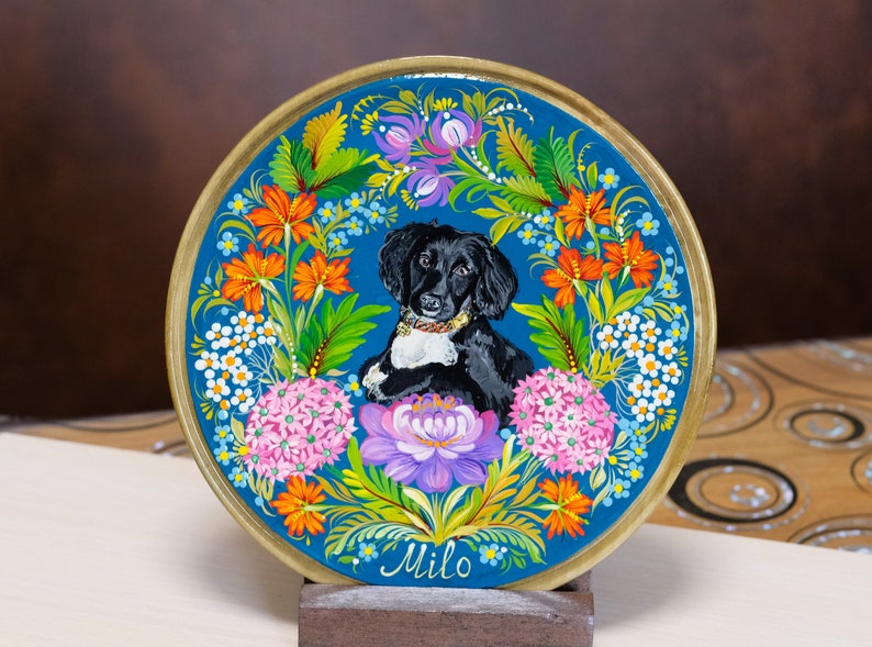 Puede incluir: Un plato de madera pintado a mano con un retrato de un perro blanco y negro rodeado de flores coloridas. El plato es azul con un borde dorado y el nombre "Milo" est&aacute; escrito en la parte inferior.