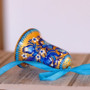 Puede incluir: Una campana dorada con dise&ntilde;os florales azules y blancos pintados en ella. La campana est&aacute; atada con una cinta azul.