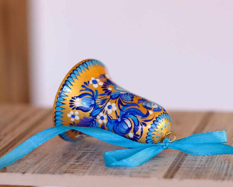 Puede incluir: Una campana dorada con dise&ntilde;os florales azules y blancos pintados en ella. La campana est&aacute; atada con una cinta azul.