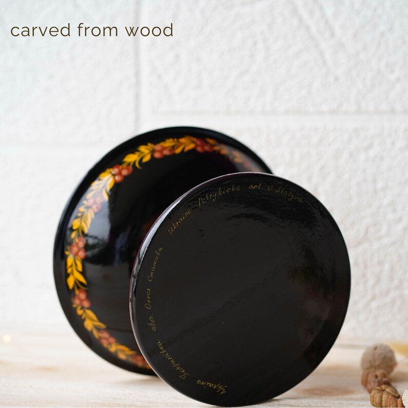 May include: A black wooden bowl with a gold and red floral design. The bowl is carved from wood and has a stand. The bottom of the bowl is inscribed with the text "ukraїns'ka  petrykivka art &  sl'stva  cel'm.  Olena  Gromula  Svitlana".
