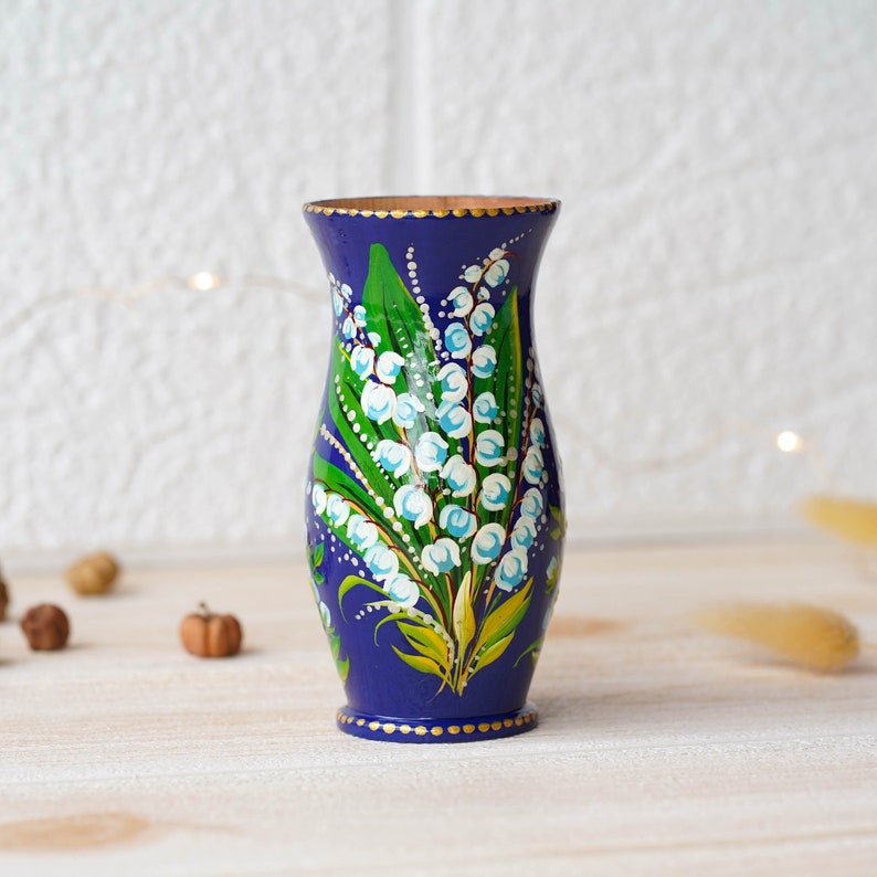 May include: A small blue ceramic vase with a hand-painted design of white lily of the valley flowers and green leaves. The vase has a gold rim and a gold dot pattern around the base.