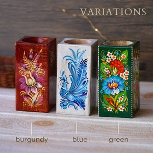 Puede incluir: Tres portavelas de madera con dise&ntilde;os pintados a mano. Los portavelas est&aacute;n pintados en burdeos, azul y verde con motivos florales y de p&aacute;jaros. Los portavelas est&aacute;n etiquetados como "burdeos", "azul" y "verde".
