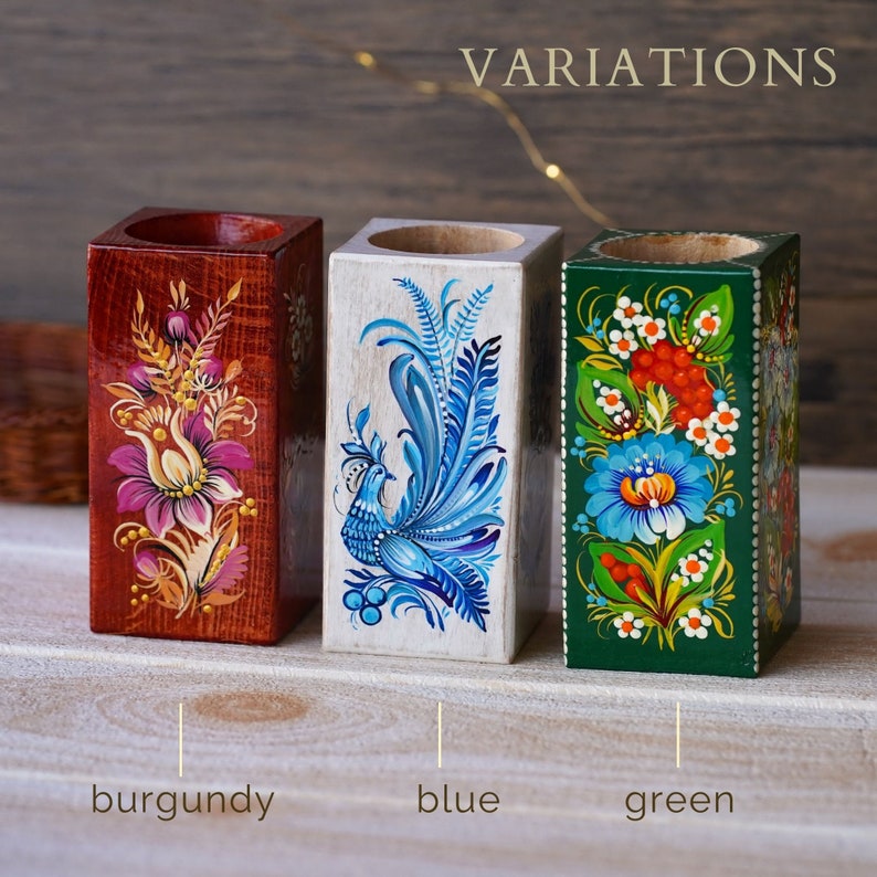 Puede incluir: Tres portavelas de madera con dise&ntilde;os pintados a mano. Los portavelas est&aacute;n pintados en burdeos, azul y verde con motivos florales y de p&aacute;jaros. Los portavelas est&aacute;n etiquetados como "burdeos", "azul" y "verde".