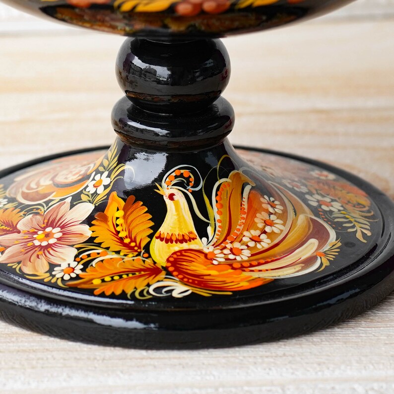 May include: A black wooden pedestal with a painted design of a bird with orange, yellow, and red feathers. The pedestal has a round base and a tall, slender column.