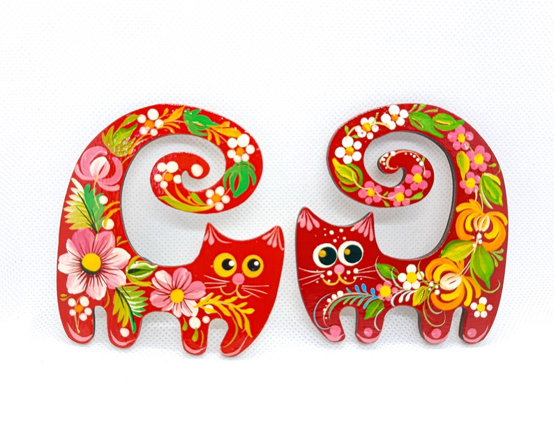 May include: Two red wooden cat figurines with curled tails. The cats are painted with colourful floral designs and have black eyes and white pupils.