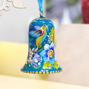 May include: A teal blue bell ornament with a painted design of a hummingbird and flowers. The bell has a gold trim and is decorated with small gold dots.
