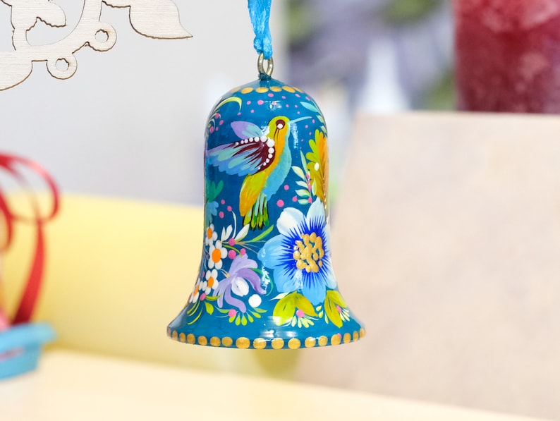 May include: A teal blue bell ornament with a painted design of a hummingbird and flowers. The bell has a gold trim and is decorated with small gold dots.