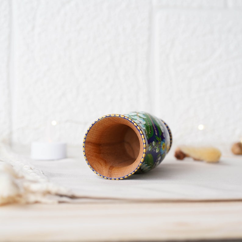 May include: A wooden cup with a blue and white floral design. The cup is painted with white flowers and green leaves on a blue background. The rim of the cup is decorated with a gold trim.
