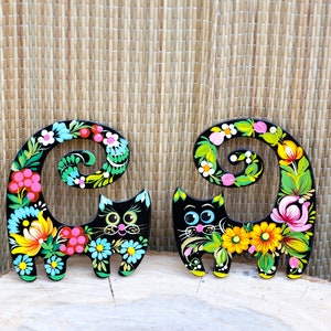 May include: Two black wooden cat figurines with curled tails painted with colorful floral designs. The cats have black eyes and pink noses.