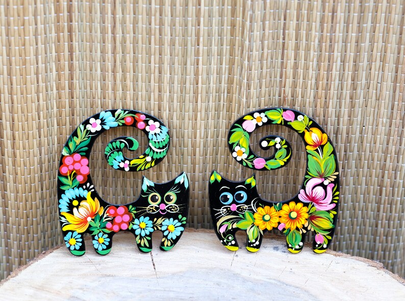 May include: Two black wooden cat figurines with curled tails painted with colorful floral designs. The cats have black eyes and pink noses.