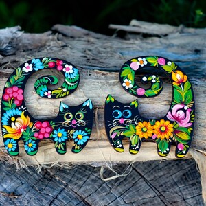 May include: Two black wooden cat figurines with colorful floral designs painted on them. The cats are curled up with their tails wrapped around their bodies. The designs feature flowers, leaves, and berries in shades of pink, yellow, blue, green, and red.