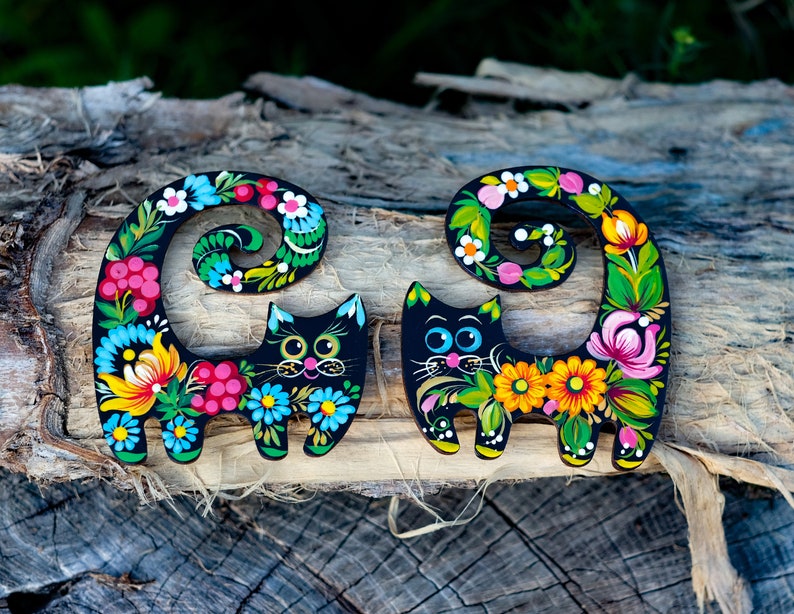 May include: Two black wooden cat figurines with colorful floral designs painted on them. The cats are curled up with their tails wrapped around their bodies. The designs feature flowers, leaves, and berries in shades of pink, yellow, blue, green, and red.