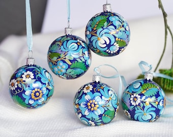 Puede incluir: Cinco adornos navideños pintados a mano en azul y blanco con diseños florales. Los adornos están colgados de una cinta blanca.