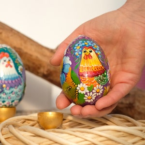 May include: A hand is holding a purple Easter egg with a painted design of a rooster and flowers. The egg is decorated with yellow, orange, green, and white paint. The word "Ukraine" is written on the egg.