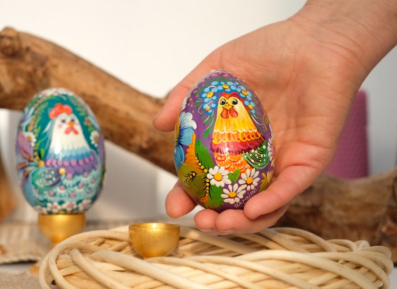 May include: A hand is holding a purple Easter egg with a painted design of a rooster and flowers. The egg is decorated with yellow, orange, green, and white paint. The word "Ukraine" is written on the egg.
