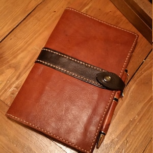 May include: A brown leather journal with a black leather strap and a gold pen. The journal has a stitched edge and a flap closure.