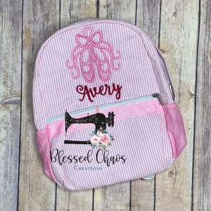 May include: Pink and white striped backpack with a pink ballet shoe design and the name "Avery" embroidered on the front. The backpack has a pink zippered pocket on the front and a mesh side pocket. The bottom of the backpack has a pink and white striped fabric with the words "Blessed Chaos Creations" embroidered on it.
