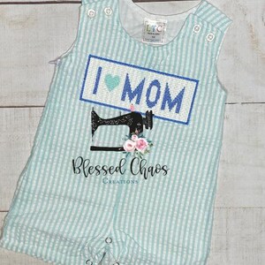 May include: A light blue and white striped baby onesie with a blue and white embroidered patch that says "I love Mom" with a heart. Below the patch is a black and white sewing machine with pink flowers. The onesie has a white text that says "Blessed Chaos Creations".