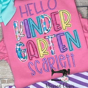 May include: Pink t-shirt with the words "HELLO KINDERGARTEN Scarlett" in colorful, embroidered letters. A large turquoise bow is at the top left. The bottom of the shirt is paired with purple and white striped pants.