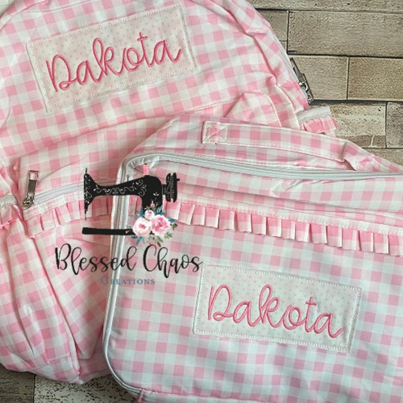 May include: Pink and white gingham backpack and lunch bag set with the name Dakota embroidered on both pieces. The lunch bag has a ruffle trim and a zipper closure.