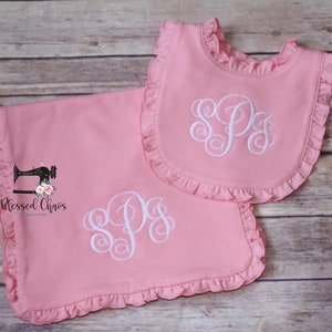 May include: A pink baby blanket and bib set with white ruffled edges and a white monogrammed "SPS" in the center. The set is made by Blessed Chaos Creations.