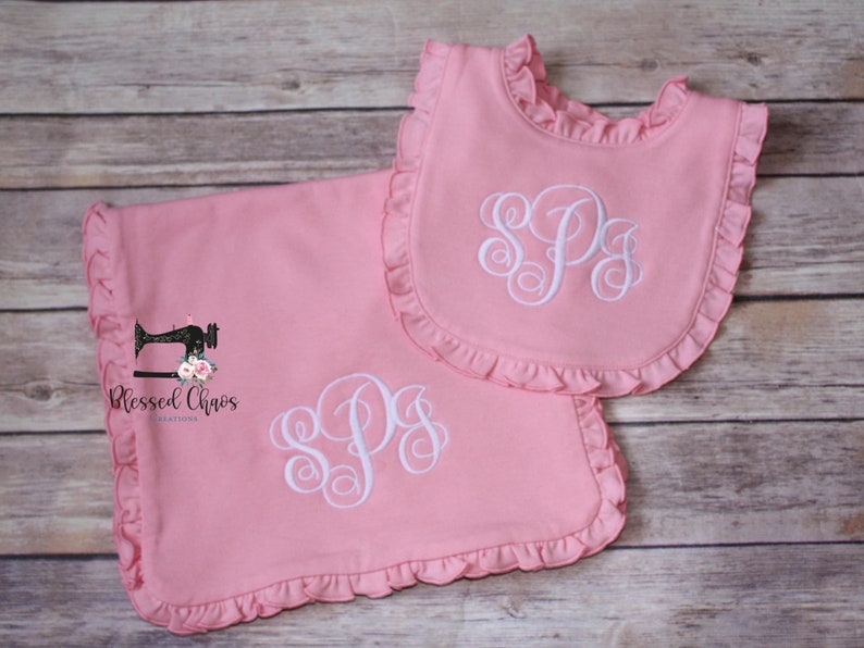 May include: A pink baby blanket and bib set with white ruffled edges and a white monogrammed "SPS" in the center. The set is made by Blessed Chaos Creations.