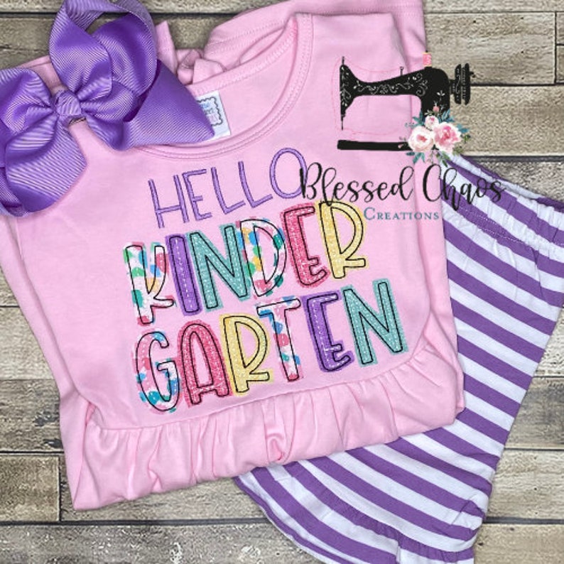 May include: A pink long-sleeved shirt and striped purple pants set. The shirt features the text "HELLO KINDERGARTEN" in colorful embroidered letters. A large purple bow is attached to the shirt. The outfit is designed for children.